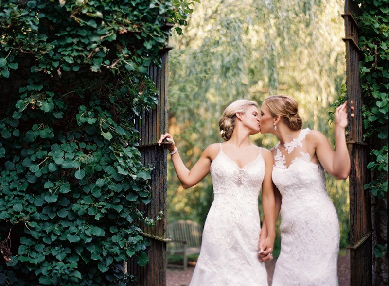 Two brides sharing a kiss at a garden gate surrounded by greenery
